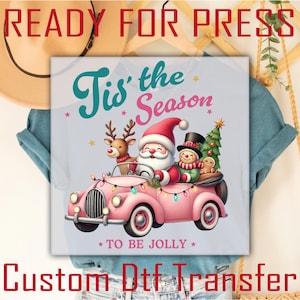 Puede incluir: Una camiseta azul claro con un diseño navideño. Muestra a Papá Noel, un reno y un muñeco de nieve en un coche rosa, con el texto "Tis' the Season To Be Jolly". La imagen está etiquetada como "Ready for Press" y "Custom Dtf Transfer".