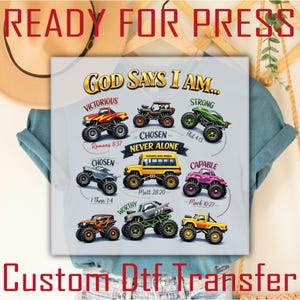 Puede incluir: Una camiseta gris claro con un gr&aacute;fico colorido con monster trucks y el texto "GOD SAYS I AM..." con palabras como "Victorioso" y "Fuerte". La camiseta tambi&eacute;n tiene el texto "READY FOR PRESS" y "Custom Dtf Transfer".