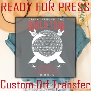 May include: Gray t-shirt with a white graphic of a geometric sphere, two hands making rock-on gestures, and the text "DRINK AROUND THE WORLD TOUR." Additional text includes locations and "ORLANDO, FL."