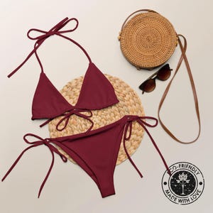 May include: A maroon bikini with a triangle top and a tie-side bottom. The bikini is on a woven straw mat with a round straw bag and sunglasses in the background. The bikini has a label that says "Eco-Friendly Made With Love".