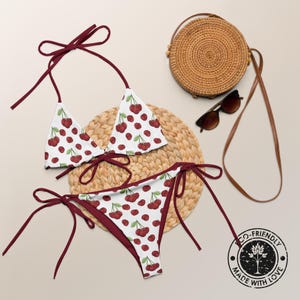 May include: A white bikini with a red cherry print and dark red tie strings. The set includes a triangle top and side-tie bottoms. A round woven straw bag, brown sunglasses, and a circular stamp reading Eco-Friendly Made With Love are visible.