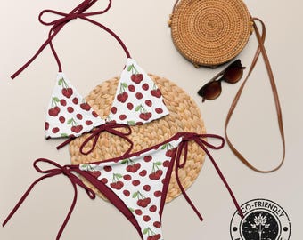 May include: A white bikini with a red cherry print and dark red tie strings. The set includes a triangle top and side-tie bottoms. A round woven straw bag, brown sunglasses, and a circular stamp reading Eco-Friendly Made With Love are visible.