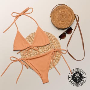 May include: A peach-colored bikini with a triangle top and a tie-side bottom. The bikini is on a woven straw mat with a round straw bag and sunglasses.