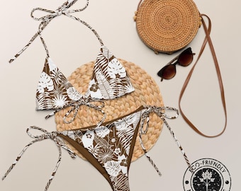 May include: A two-piece bikini set featuring a brown and white tropical leaf and floral pattern with adjustable side ties on the bottoms and a halter-style top. Included are a round woven straw crossbody bag and dark brown sunglasses.