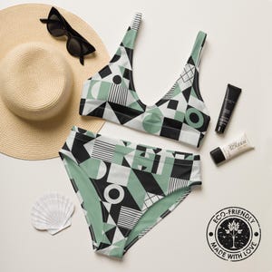 May include: A two-piece bikini set featuring a geometric pattern in sage green, black, and white. The set includes a scoop-neck top and high-waisted bottoms. Also shown are a straw hat, black sunglasses, two tubes of sunscreen, and a seashell.