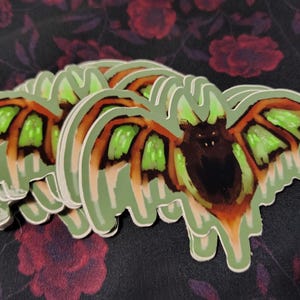 May include: A stack of die-cut stickers featuring a stylized bat with a dark brown body, orange wing accents, and bright neon green wing membranes. The bat has pointed ears and a small face, with a light green border outlining the entire shape.