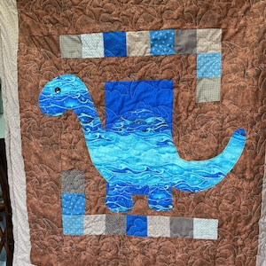 May include: A handmade quilt featuring a blue dinosaur with a long neck and tail, set against a brown background. The dinosaur has a water-like pattern. The quilt has a patchwork border with squares of blue, grey, and white fabrics.