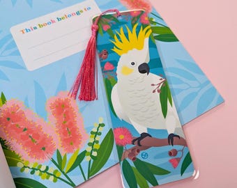 May include: A colourful bookmark featuring a white cockatoo with a yellow crest perched on a branch with green leaves and pink flowers. The bookmark has a pink tassel.