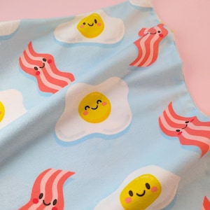 May include: Pale blue fabric featuring a repeating pattern of cartoon fried eggs and bacon rashers. The eggs have yellow yolks and smiling faces. The bacon rashers are pink and red with smiling faces. Suitable for kitchen or breakfast-themed products.