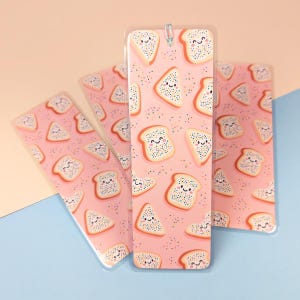 May include: Four clear plastic bookmarks featuring a pink background and a repeating pattern of smiling toast slices with sprinkles. A silver paper clip secures one bookmark. The background is a gradient of pink and blue.