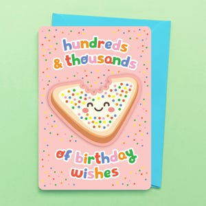 May include: A pink birthday greeting card featuring a cartoon slice of bread covered in colorful sprinkles with a smiling face. The text reads hundreds & thousands of birthday wishes. The card is paired with a bright blue envelope.