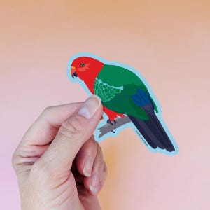 以下が含まれることがあります： 鮮やかなキングパロットのステッカー。鳥は鮮やかな赤い頭、緑の翼、青い尾を持っています。明るいピンク色の背景に、枝にとまっています。ステッカーは手で持たれています。