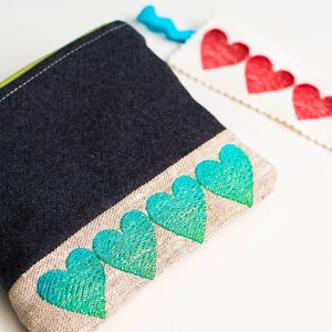 May include: A black fabric pouch with a green zipper and a row of turquoise embroidered hearts along the bottom edge. A white fabric strip with red embroidered hearts is also shown.