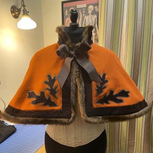 May include: A brown and orange cape with a faux fur trim and black velvet accents. The cape features a large black bow and two embroidered oak leaf designs.