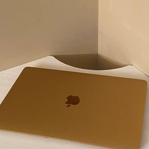 Puede incluir: Una estanter&iacute;a de esquina blanca con un borde frontal curvado. Un ordenador port&aacute;til dorado cerrado con el logotipo de Apple est&aacute; colocado en la estanter&iacute;a. La estanter&iacute;a est&aacute; montada en una pared beige.
