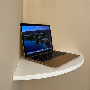 Puede incluir: Un ordenador port&aacute;til dorado con un teclado negro est&aacute; abierto sobre una estanter&iacute;a blanca y curvada. La pantalla muestra una imagen panor&aacute;mica de un lago y monta&ntilde;as. El port&aacute;til est&aacute; colocado en una esquina contra una pared beige.