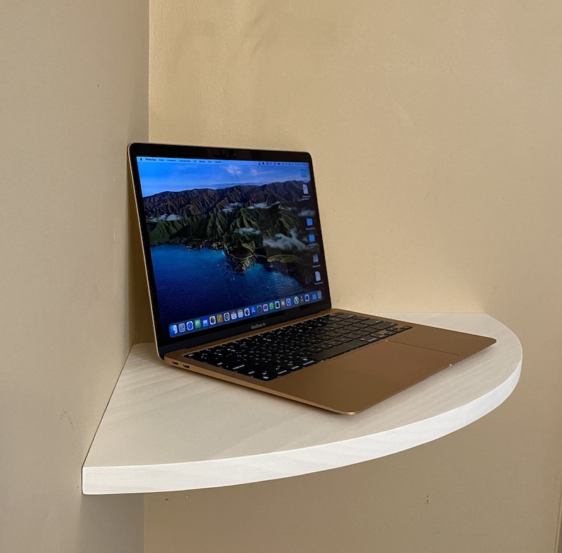 Puede incluir: Un ordenador port&aacute;til dorado con un teclado negro est&aacute; abierto sobre una estanter&iacute;a blanca y curvada. La pantalla muestra una imagen panor&aacute;mica de un lago y monta&ntilde;as. El port&aacute;til est&aacute; colocado en una esquina contra una pared beige.