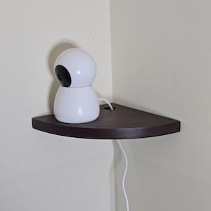 May include: A white security camera with a black lens sits on a dark brown, triangular corner shelf. The shelf is mounted on a light beige wall. A white power cord extends from the camera.