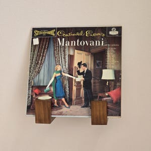 May include: A vintage vinyl record album cover for Mantovani and his orchestra, mounted on a wooden display. The album cover features a retro illustration of a couple in a room. The album is held in place by two wooden supports.