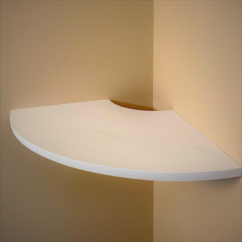 Puede incluir: Estanter&iacute;a de esquina blanca y semicircular montada en una pared beige. La estanter&iacute;a tiene un borde frontal curvo y est&aacute; dise&ntilde;ada para encajar perfectamente en una esquina. La estanter&iacute;a es de madera y est&aacute; pintada de blanco.