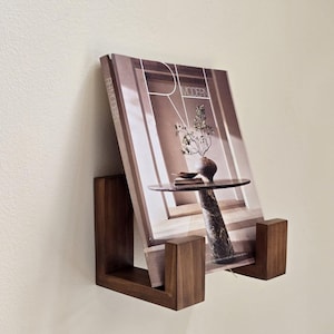 Peut inclure: Un porte-revues en bois brun foncé fixé au mur. Il contient un magazine avec le texte "RH MODERN" visible. Le magazine présente une photographie d'une table avec un vase et une plante.