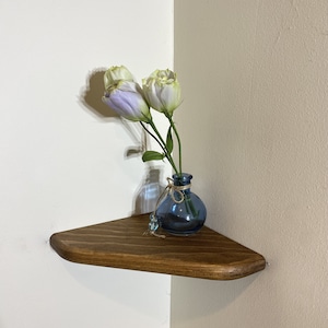 May include: A dark brown, triangular corner shelf with a rounded edge. A small, blue glass vase with a twine tie holds two white and purple flowers. The shelf is mounted on a white and beige wall.