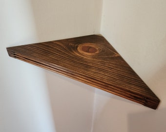 May include: A triangular wooden shelf with a dark brown stain. The shelf is mounted in a corner of a room.