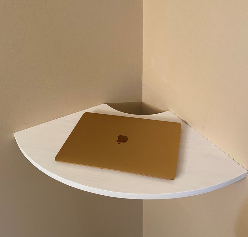Puede incluir: Una estanter&iacute;a de esquina blanca con un borde frontal curvado. Un ordenador port&aacute;til dorado cerrado con el logotipo de Apple est&aacute; colocado en la estanter&iacute;a. La estanter&iacute;a est&aacute; montada en una pared beige.