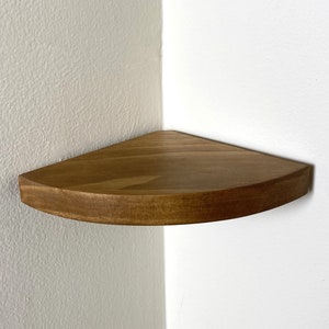 May include: A wooden corner shelf with a rounded edge. The shelf is made of light brown wood and is mounted on a white wall.