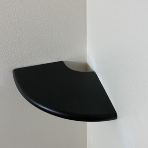 Puede incluir: Un estante de esquina negro montado en una pared blanca. El estante está curvado para adaptarse a la esquina.