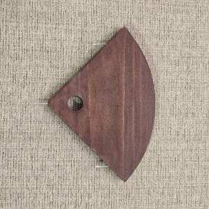 May include: A dark brown, fan-shaped wooden serving tray with a small circular hole and two thin metal rods. The tray has a smooth finish and is set against a textured, neutral-colored background.
