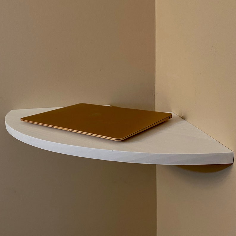 Puede incluir: Una estanter&iacute;a de esquina blanca, semicircular, con un port&aacute;til dorado encima. La estanter&iacute;a est&aacute; montada en una pared beige, creando un dise&ntilde;o minimalista y funcional. El port&aacute;til est&aacute; cerrado y es dorado.