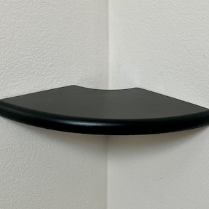 May include: A black corner shelf with a curved front edge. The shelf is mounted on a white wall. The shelf is designed to fit into a corner, maximizing space.