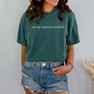 May include: A teal green t-shirt with the phrase "I'm my husband's problem" printed in white. The shirt is paired with denim shorts. The model is wearing a necklace and bracelets.