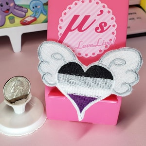 May include: A heart-shaped embroidered patch with wings, featuring the asexual pride flag colors: black, white, and purple. The patch is on a pink background with a white star-shaped border and the text "μ's Love Live!"