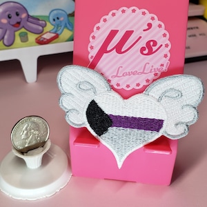 May include: A white heart with black and purple stripes, with wings, is embroidered on a pink background. The heart has a white outline and the wings are silver. The background has the text "μ's Love Live!" in pink and white.