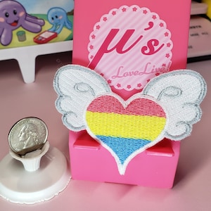May include: A white heart with pink, yellow, and blue stripes, with white wings, and the text "Love Live!" on a pink background.