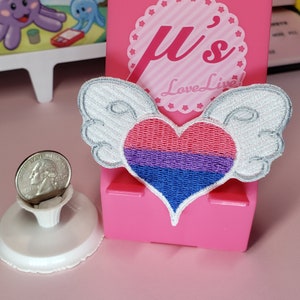 May include: A white heart with wings embroidered with the bisexual pride flag colors. The heart is pink, purple, and blue. The wings are white with a silver outline. The heart is on a pink background with the text "μ's Love Live!" in white.