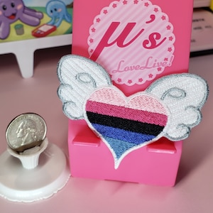 May include: A white heart with wings embroidered patch with a pink, red, black, blue, and light blue striped heart. The patch is on a pink phone stand with the text "μ's LoveLive!" on it.