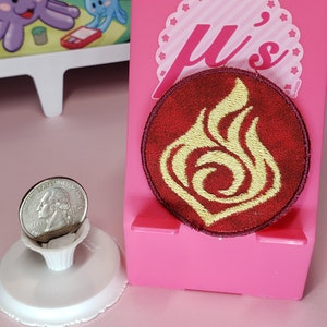 May include: A red and gold embroidered patch with a stylized flame design. The patch is circular and has a white border. The text "Ju's" is visible in the background.