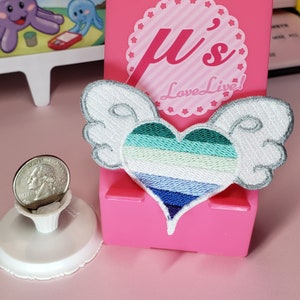 May include: Embroidered patch with a heart design, featuring layered stripes in blue, green, and white, and white wings. The patch is set against a pink background. The patch is a decorative item.