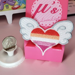 May include: A white heart with rainbow stripes and wings, with the text "Love Live!" on a pink background.