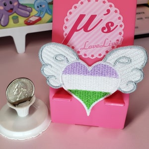 May include: Embroidered patch featuring a heart with genderqueer flag colors, white wings, and a silver outline. The patch is displayed on a pink stand with the text "μ's Love Live!". A quarter coin is in the foreground.