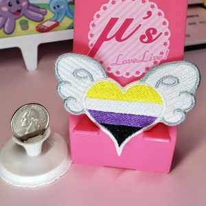 May include: A white heart with wings, featuring the colors of the non-binary pride flag, yellow, white, purple, and black. The heart is embroidered and has a white outline. The heart is on a pink background with the text "μ's Love Live!" in white.