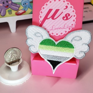 May include: A green, white, and black heart-shaped patch with wings. The heart is a representation of the genderqueer pride flag. The patch is attached to a pink phone stand with the text "μ's Love Live!" on it.