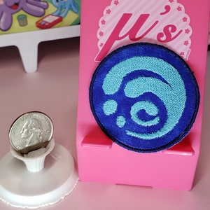May include: Blue and teal embroidered patch with a swirling design. The patch is circular and has a white background. The patch is attached to a pink stand.