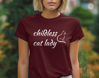 Peut inclure: Un t-shirt bordeaux avec le texte "childless cat lady" en écriture cursive blanche et une silhouette blanche d'un chat assis sur ses haunches.