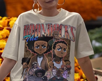 May include: Beige t-shirt featuring a graphic from "The Boondocks" animated series. The design includes characters from the show and the show's logo. The person in the image is wearing black pants and gold hoop earrings.