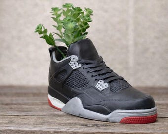 May include: A black and gray sneaker-shaped planter with a red sole, holding a small green plant. The planter is made to resemble a popular athletic shoe design. The sneaker planter sits on a wooden surface.