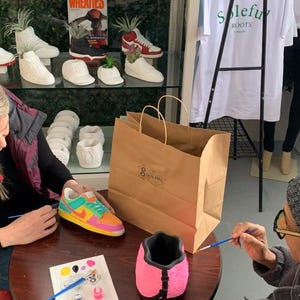 May include: A craft store display featuring painted sneaker-shaped planters and a variety of other decorative planters. A person is painting a pink planter. A colorful sneaker planter is on the table. A brown paper shopping bag is on the table.